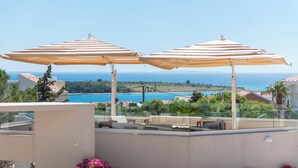 A terrace with two beige umbrellas, a stone wall, and a view of the sea and buildings. - Apartments Dyana (Novalja)
