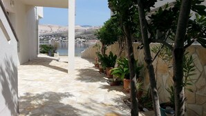 A stone-paved patio with potted plants and a view of a lake and buildings in the distance. - Apartments Ena (Pag)