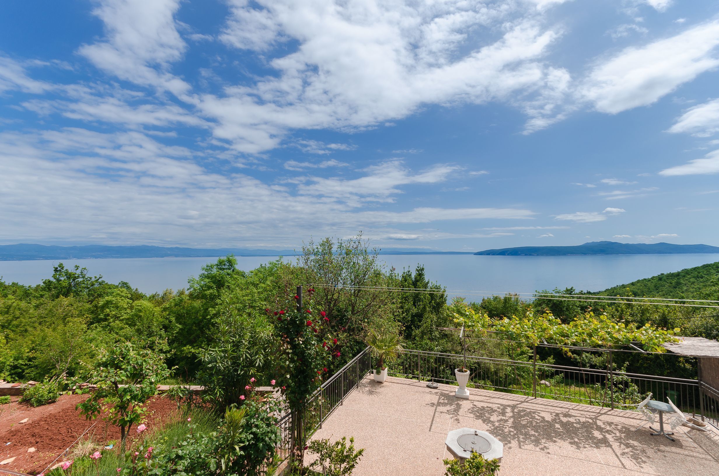 A terrace with a view of a lake and mountains, surrounded by greenery and potted plants.