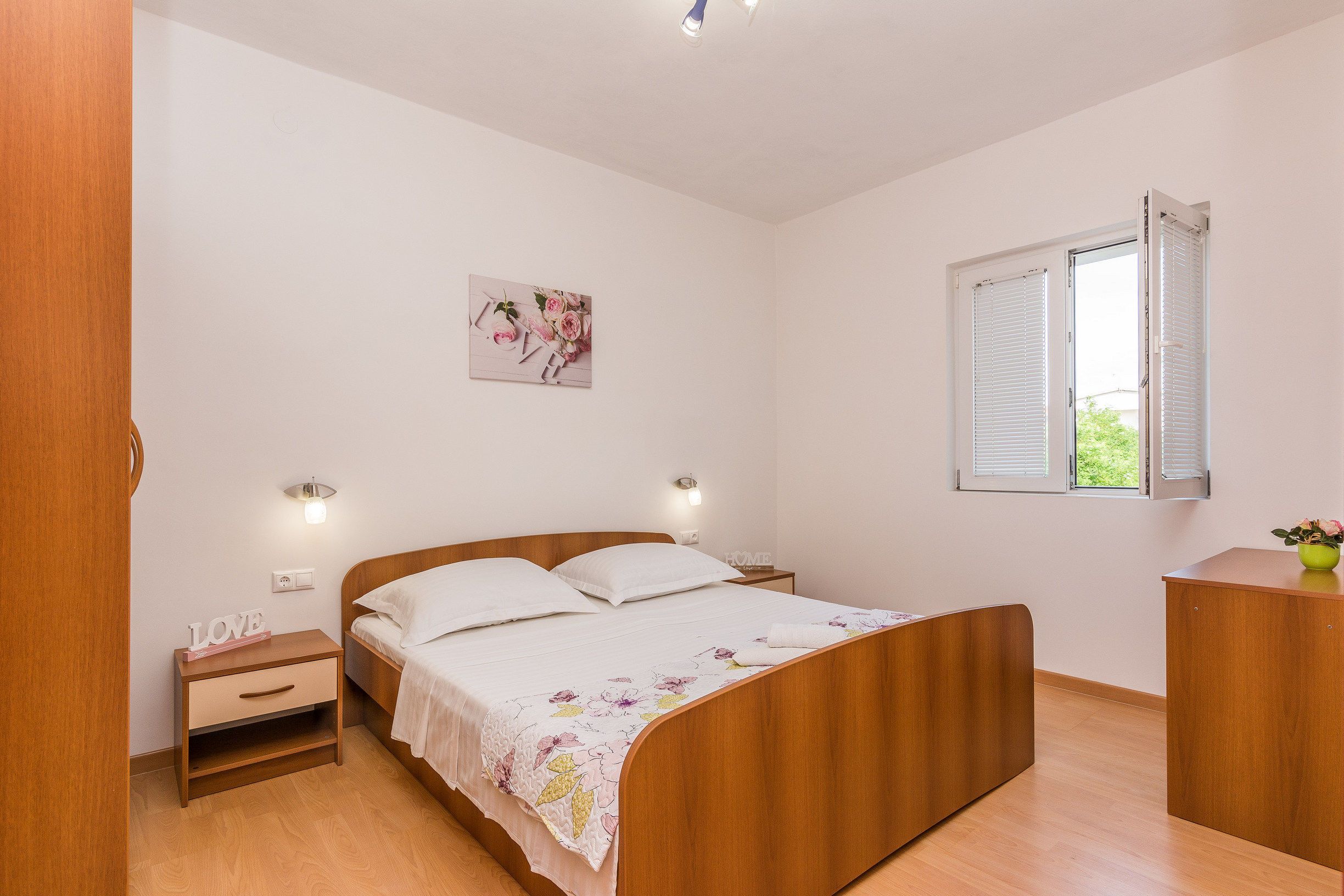 Een slaapkamer met een houten bed, nachtkastjes, een wandlamp, een raam met luiken en een bloemrijk kunstwerk.