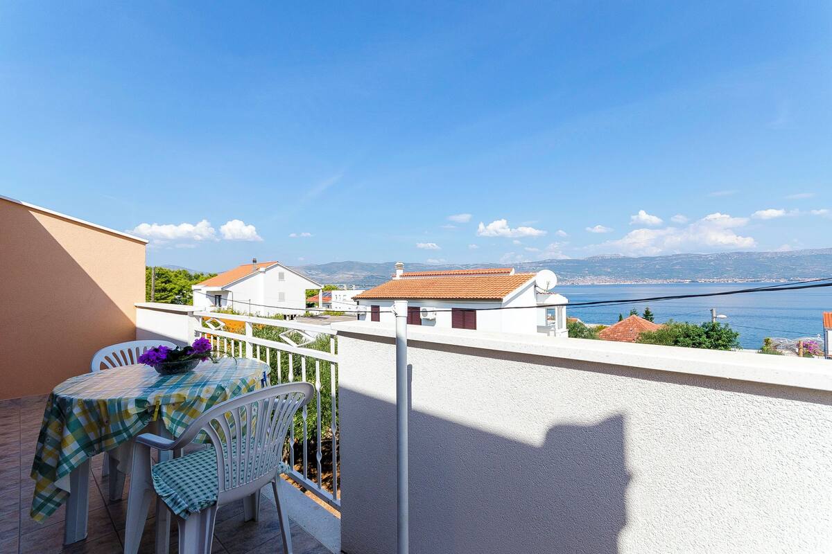 A balcony with a table and chairs, overlooking a coastal view with houses and a clear sky.