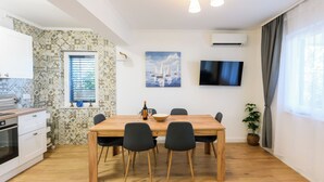A modern kitchen with white cabinets, a wooden dining table with four chairs, a TV mounted on the wall, and a window with curtains. - Apartments Milka (Slatine)