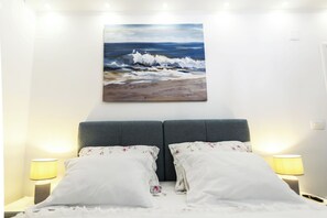 A bedroom with a bed, two pillows, a nightstand with a lamp, and a painting of a beach scene on the wall.