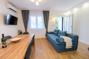 A modern living room with a wooden dining table, a blue sofa, and a flat-screen TV mounted on the wall.