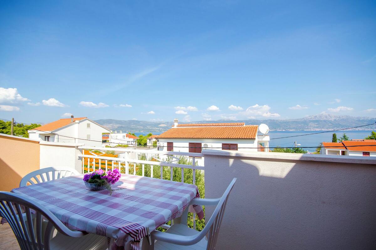 A balcony with a table set for two, overlooking a coastal town with houses and mountains in the distance.