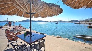 A beachside table with a straw umbrella, a chair, and a bottle. - Apartments Smilja (Tribunj)