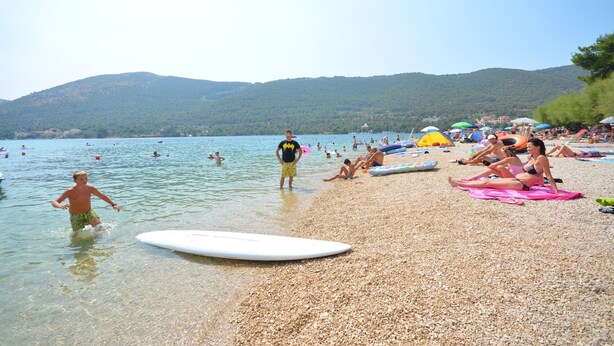 Een kiezelstrand met mensen die zwemmen en zonnebaden, een surfplank en parasols.
