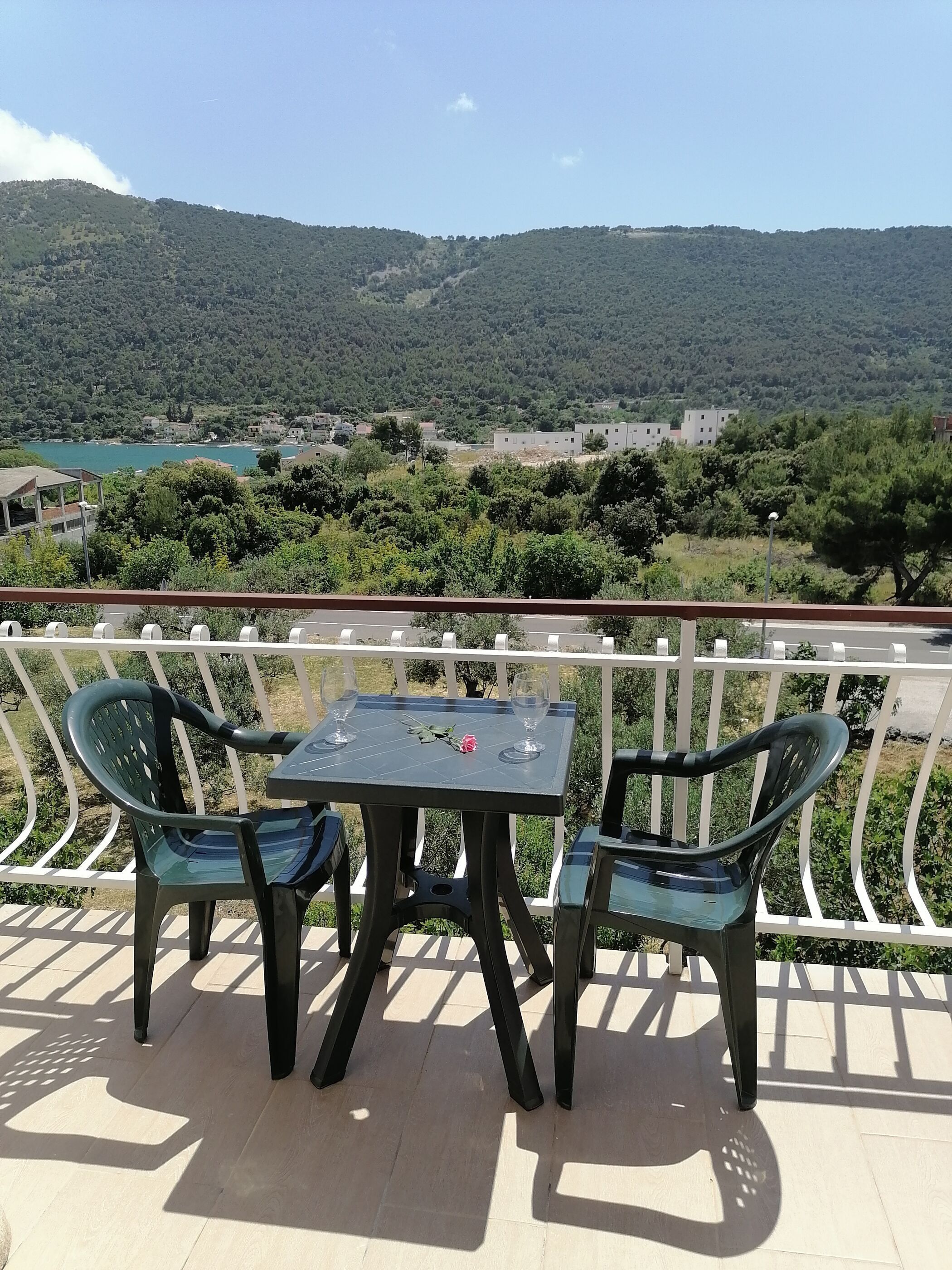 A balcony with two chairs and a table, overlooking a scenic view of a hilly landscape with buildings and a body of water.
