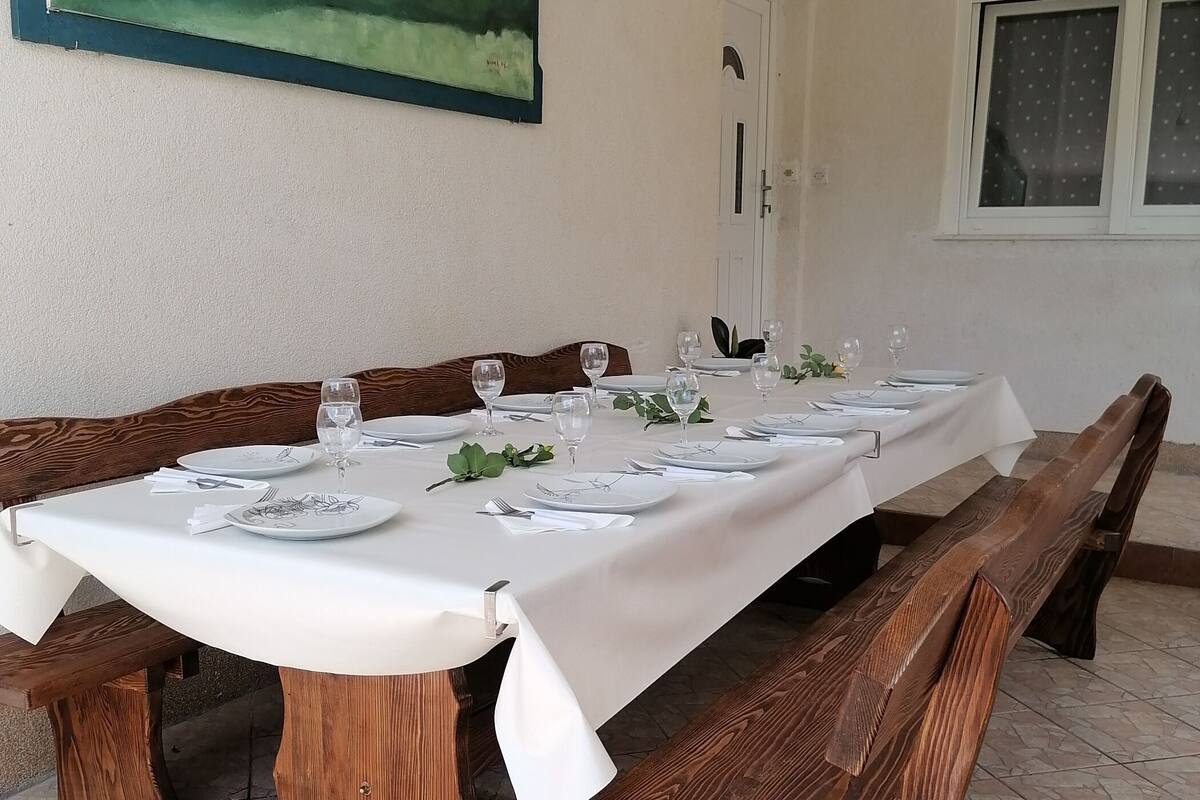 A dining table set for six with white tablecloths, wooden benches, and a painting on the wall.