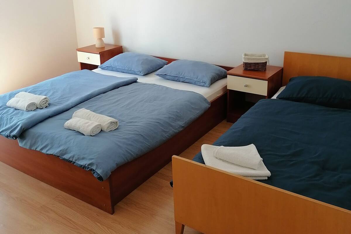 A double bed with blue bedding and two pillows, a bedside table with a lamp, and a small bedside table with a basket.