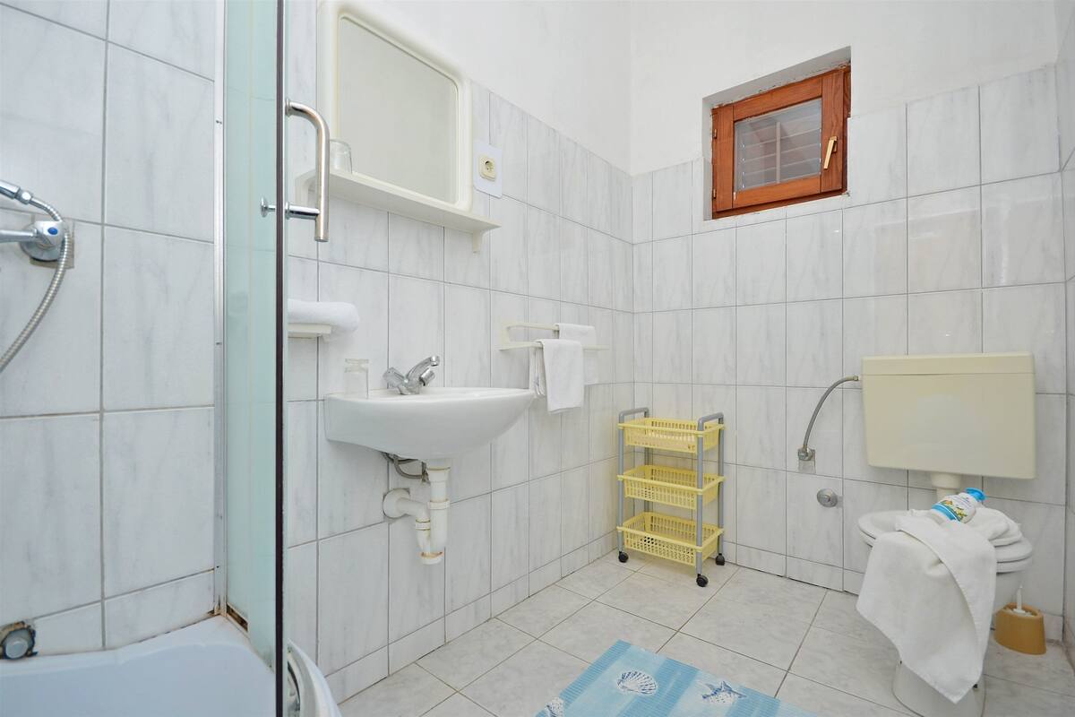Egy fürdőszoba, amelyben mosdókagyló, WC, zuhanyzó és egy kis ablak található.