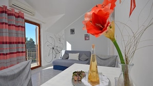 A modern living room with a grey sofa, a dining table with a vase of flowers, and a large window with curtains.