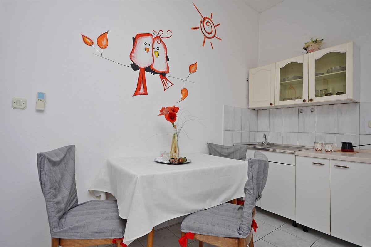 A dining area with a table set for two, chairs, a vase with flowers, and a wall with a bird mural.