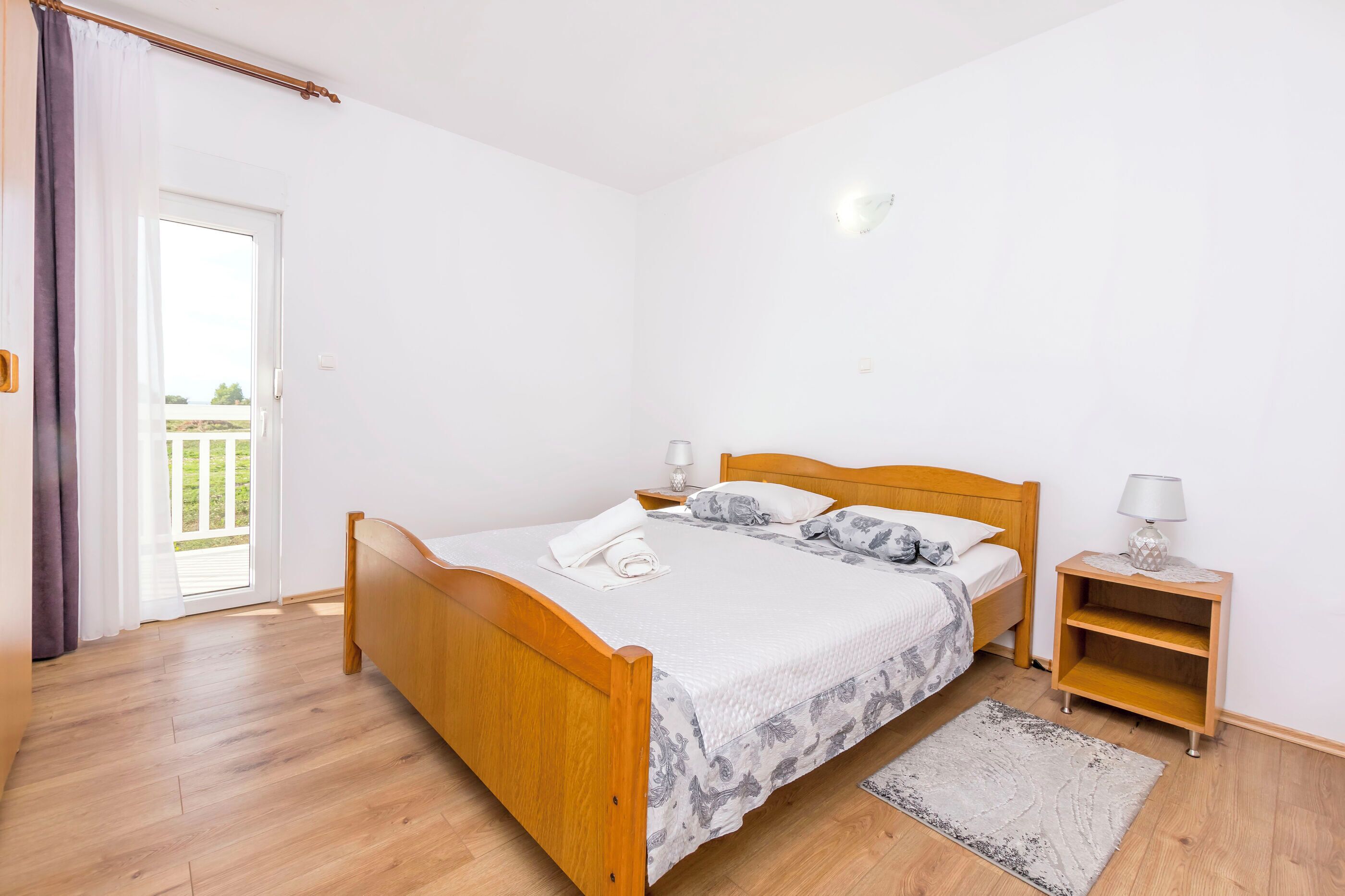 A bedroom with a wooden bed, a nightstand with a lamp, a small wooden shelf, and a view of the outdoors through a sliding door.