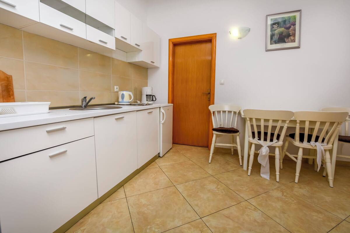A kitchen with white cabinets, a sink, and a coffee maker. There is a wooden door and a framed picture on the wall.