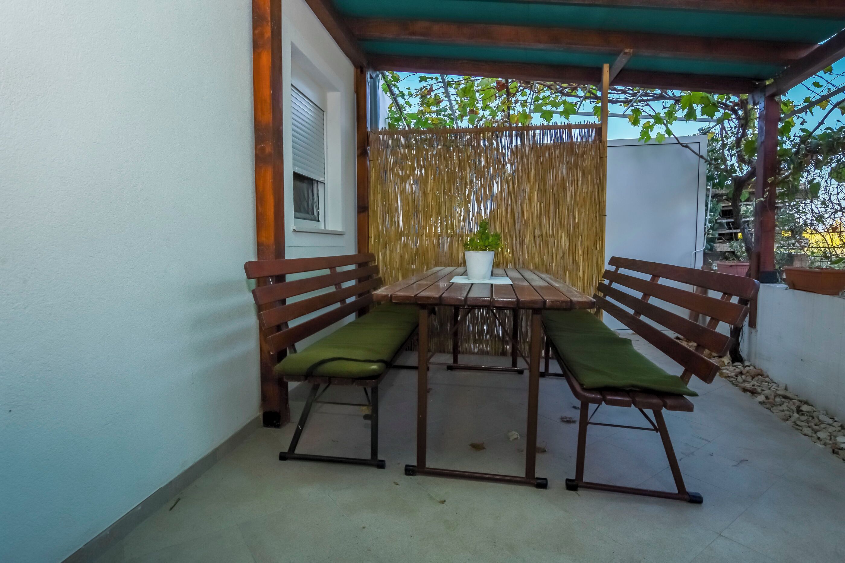 A covered patio area with a wooden table and two benches, each with green cushions.