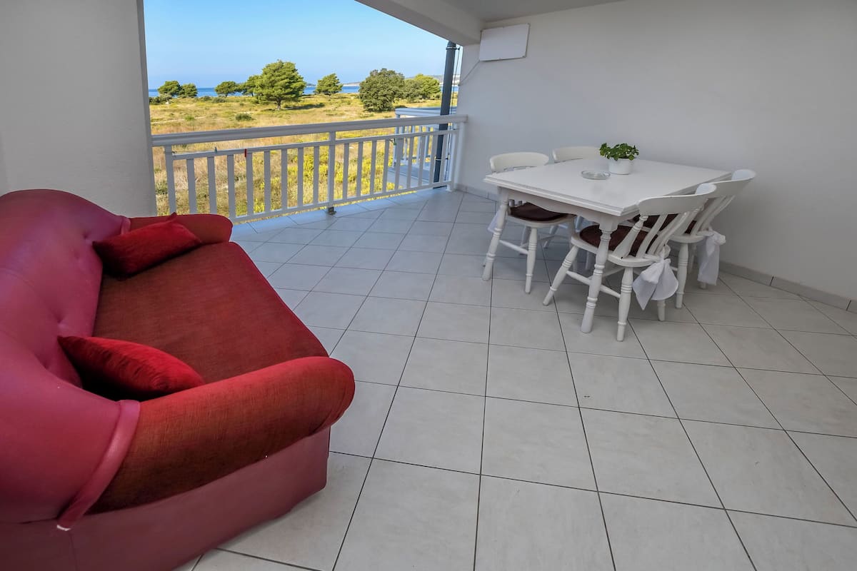 A balcony with a white table and chairs, a red sofa, and a view of the sea and trees.