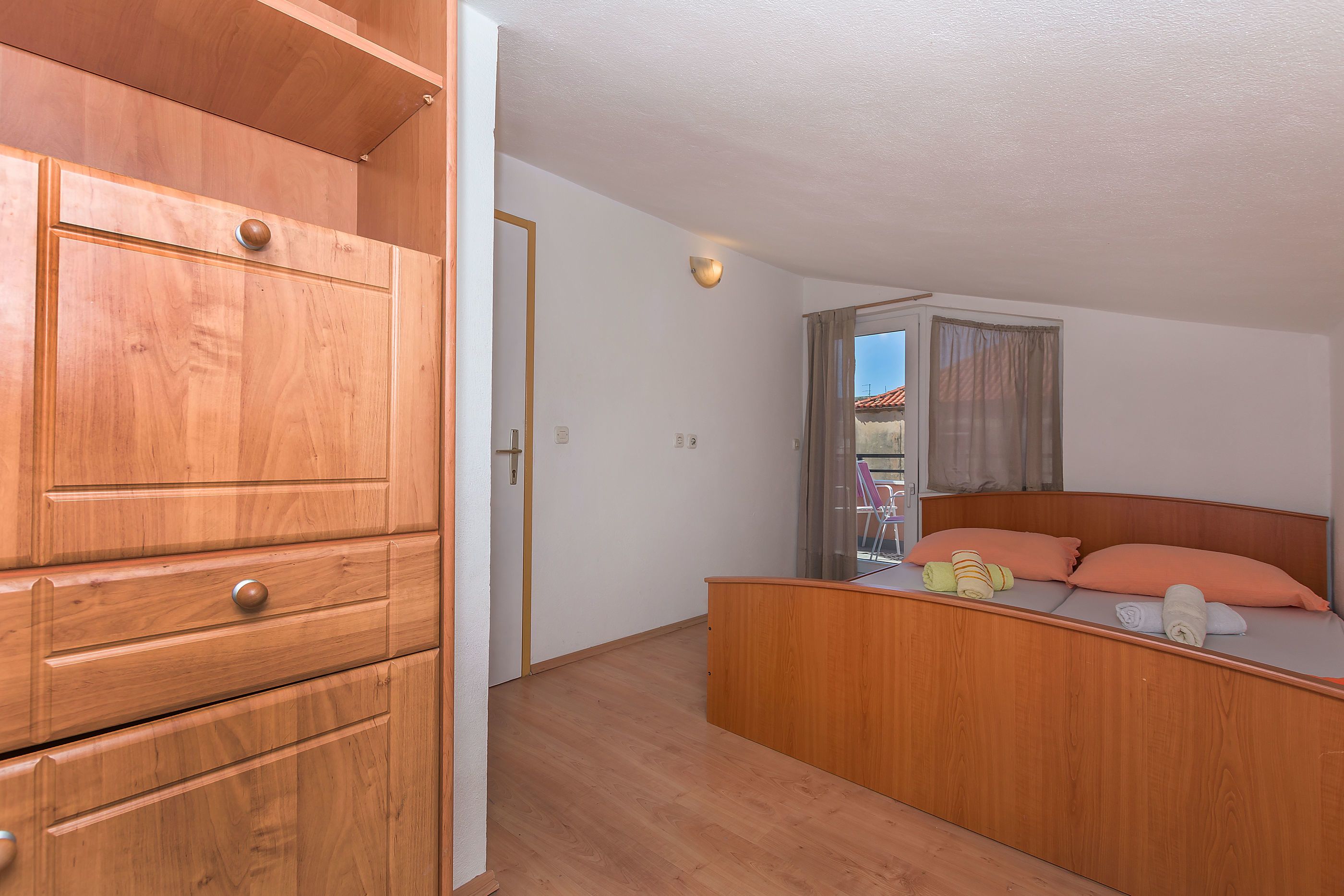 Un armario de madera con varios cajones y una cama con cabecero, una ventana con cortinas, y un pequeño balcón visible a través de la ventana.