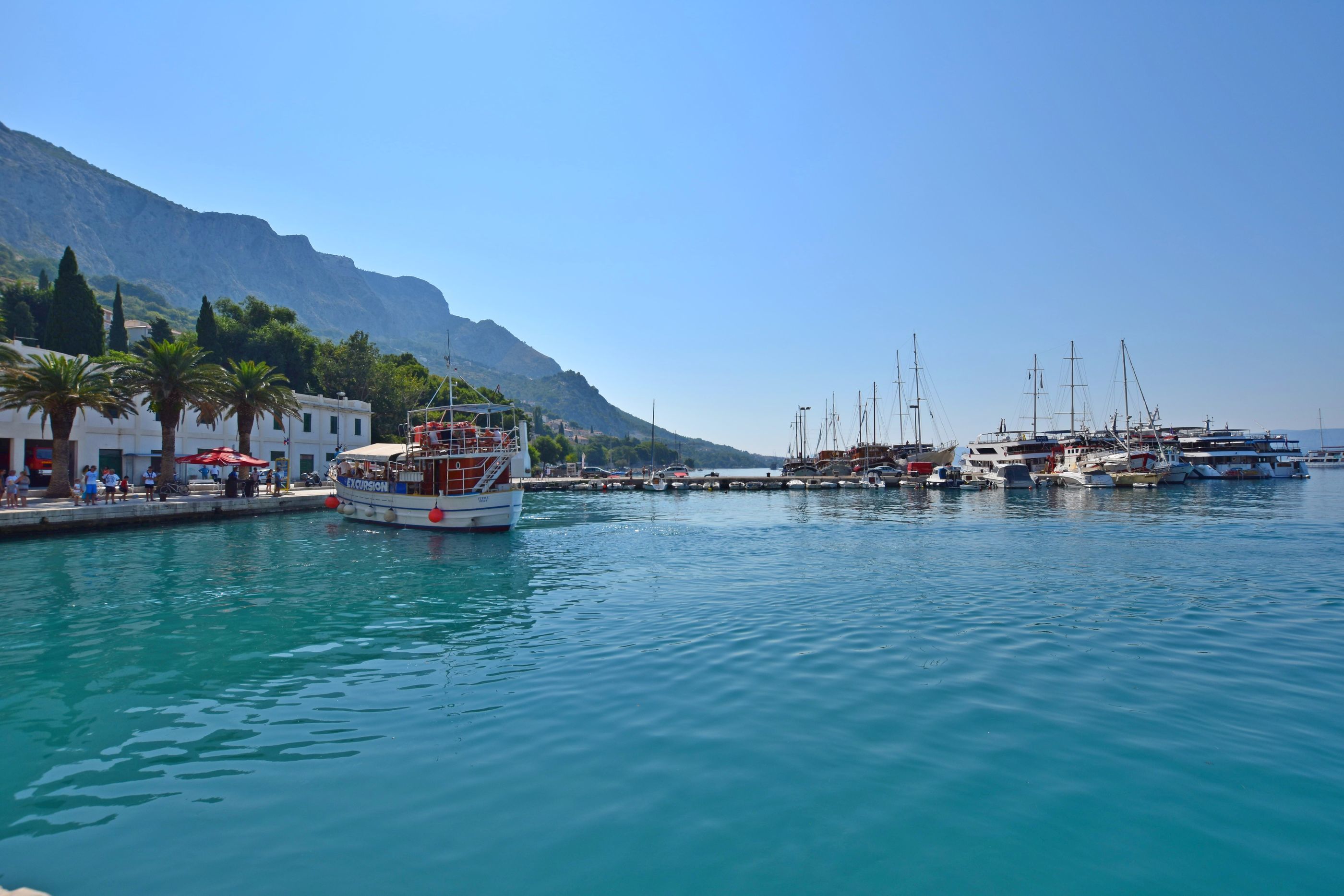A marina with boats docked, a white building, and a mountain in the background.