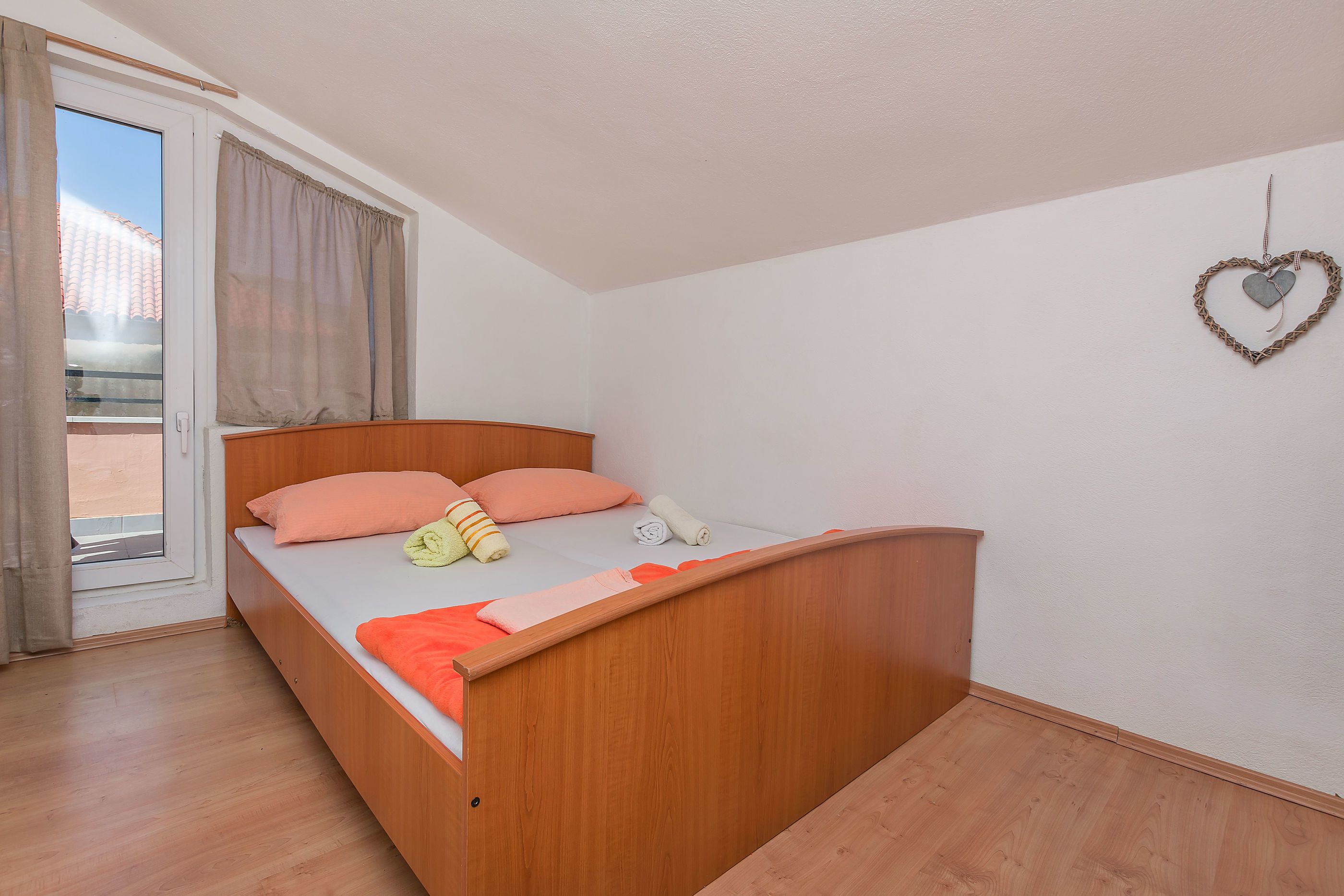 A single bed with a wooden frame, a window with curtains, and a heart-shaped decoration on the wall.