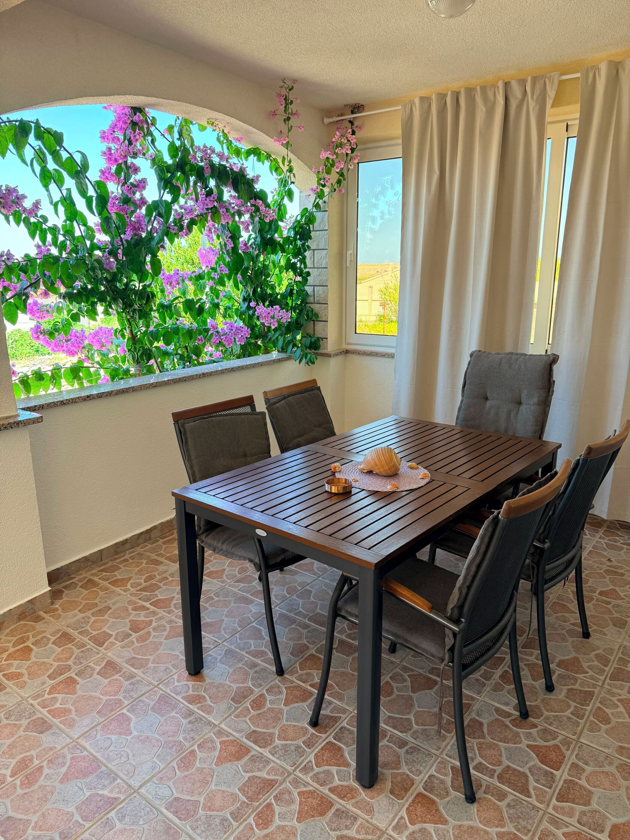 Ein Essbereich mit Tisch, Stühlen und Blick auf violette Blumen durch ein Fenster.