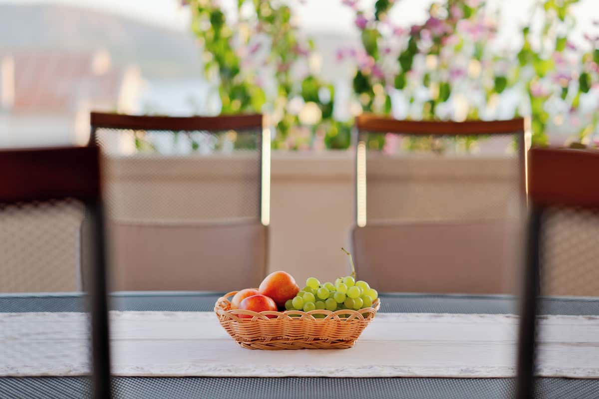 A wicker fruit basket on a table with a lace tablecloth, two wooden chairs, and a blurred floral background.