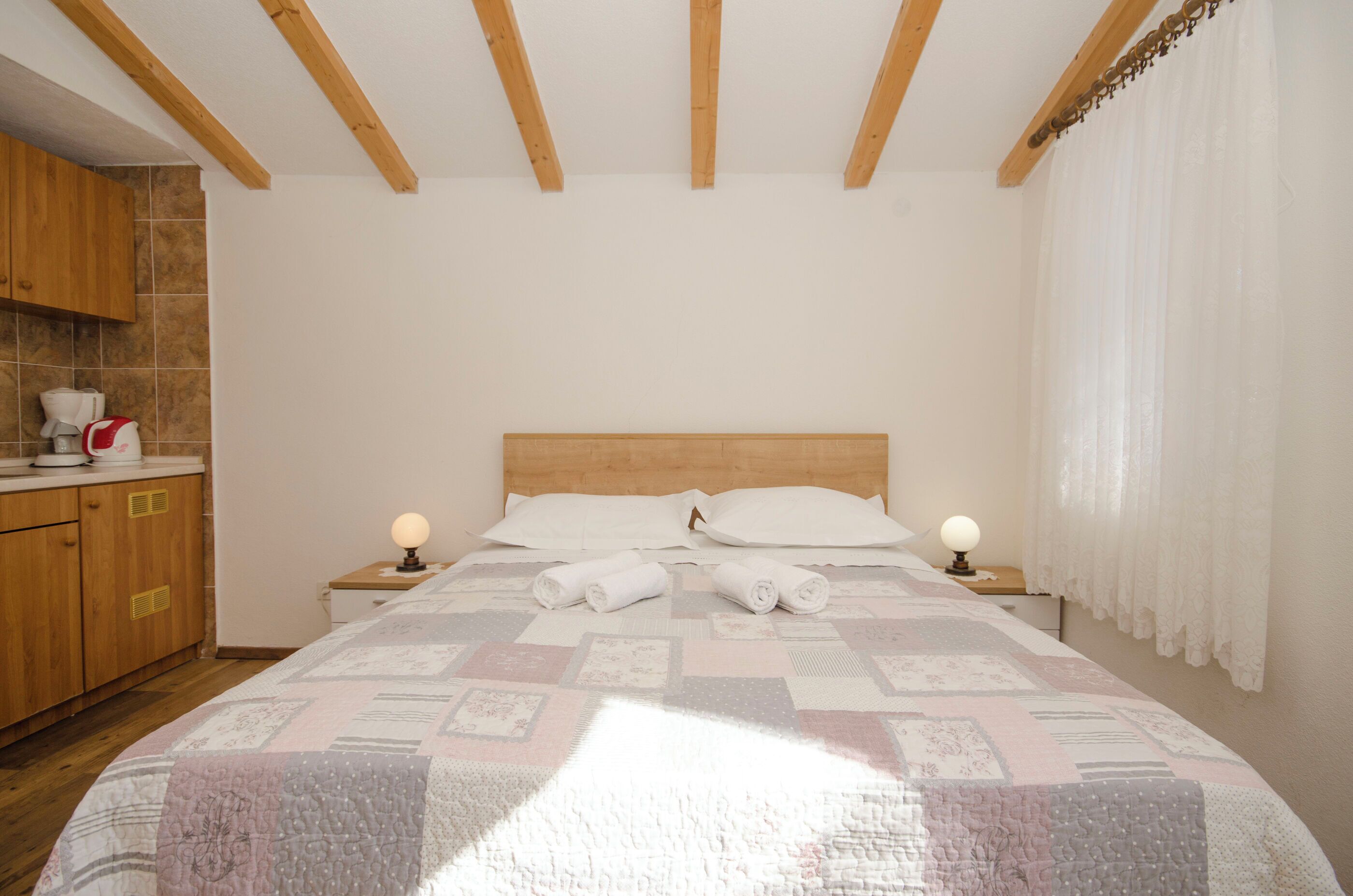 A bedroom with a wooden headboard, a quilted bedspread, two bedside lamps, and a wooden cabinet with a kettle.