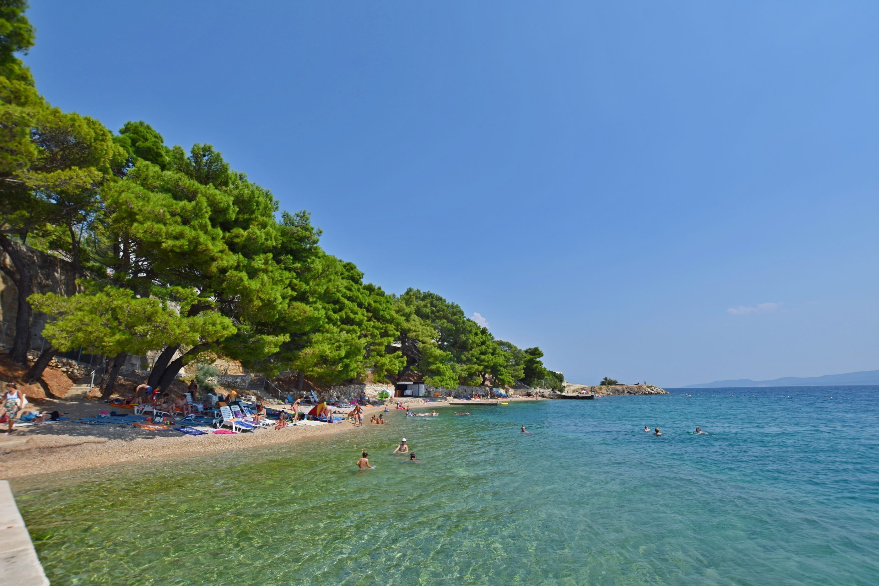A beach with clear water, people swimming and sunbathing, and trees along the shore.