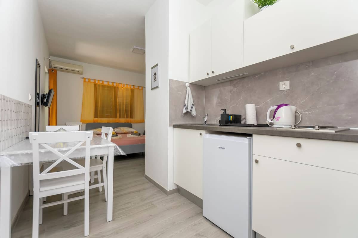 A compact kitchen with a white refrigerator, a coffee maker, and a sink. There is a small dining table with chairs and a bed in the background.