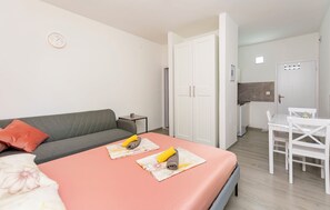 Een moderne slaapkamer met een bed, een grijze bank, een klein tafeltje met een stoel, en een kitchenette op de achtergrond.