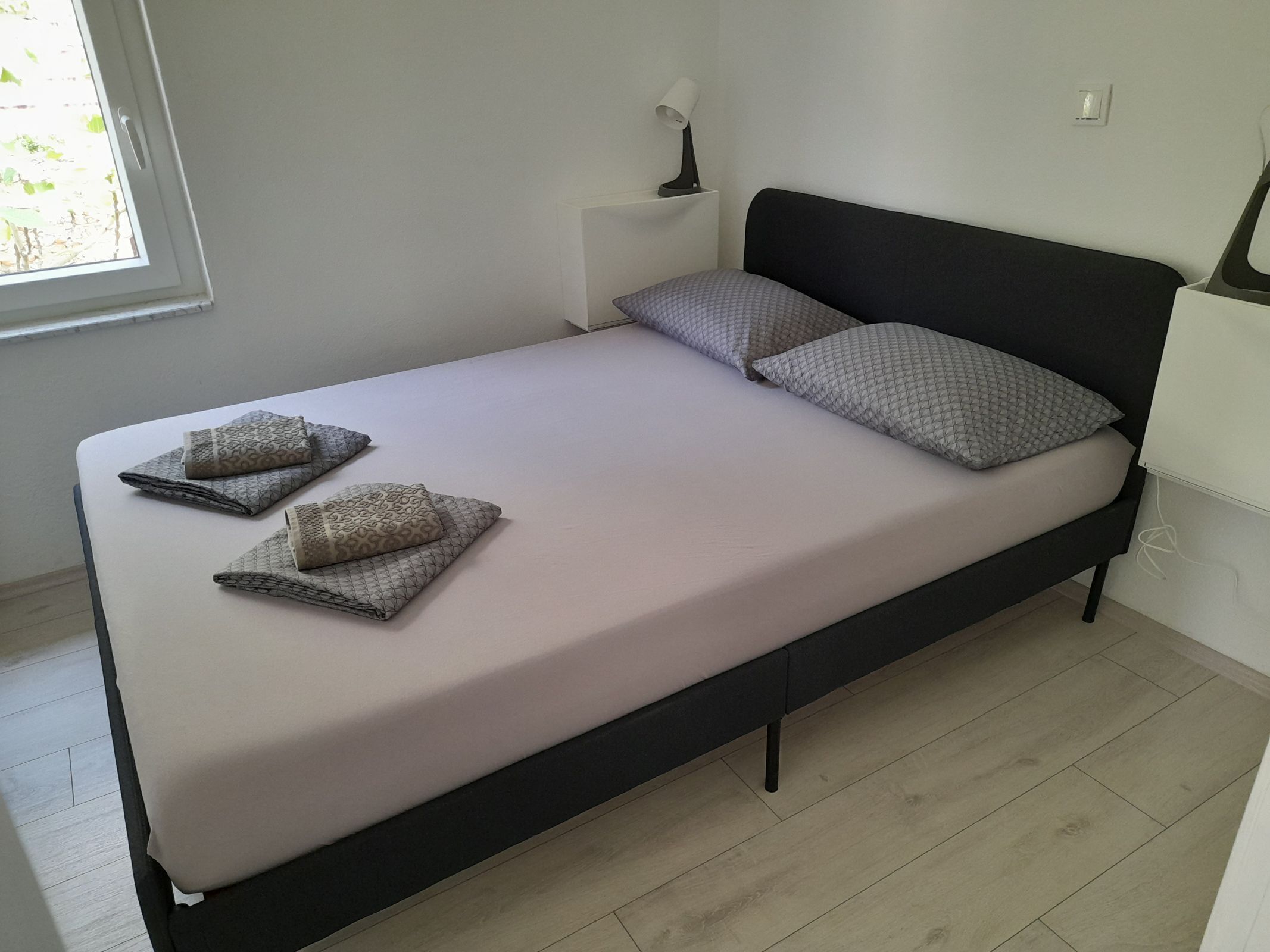 A neatly made bed with pillows and folded blankets, a bedside lamp, and a window with blinds.