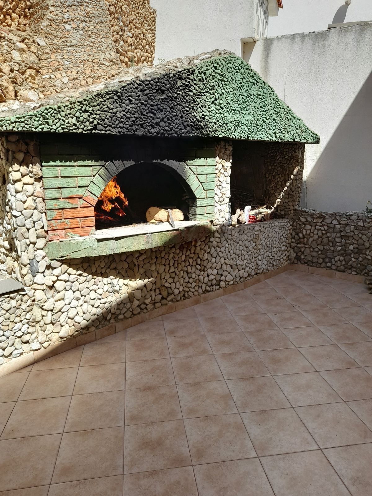 A stone-built outdoor pizza oven with a green roof, surrounded by a stone wall and a tiled patio area.