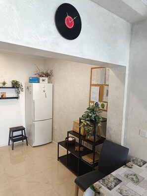 A compact living space with a refrigerator, a small table, a chair, a black clock, and a bulletin board with papers.