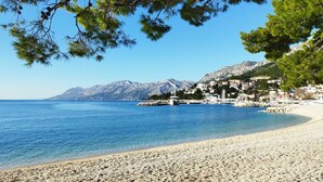 Ein Kiesstrand mit klarem, blauem Meer, eine Stadt am gegenüberliegenden Ufer und Berge im Hintergrund.