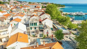 A coastal town with terracotta-roofed buildings, a marina with boats, and a promenade with outdoor seating.