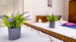 A bedroom with a wooden headboard, a bedside table with a lamp, and two potted plants with purple flowers.