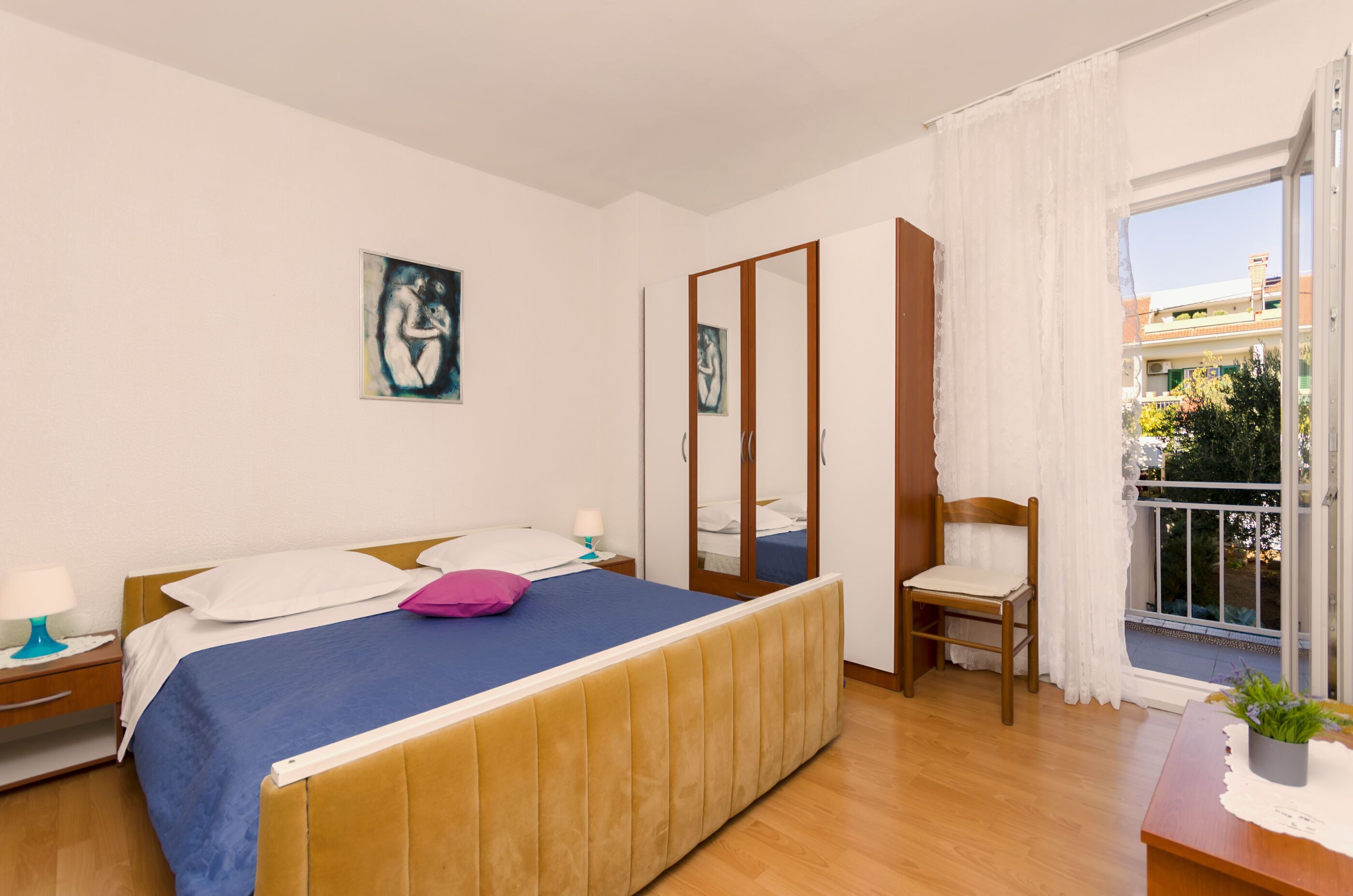 A bedroom with a bed, a chair, a nightstand, a window with a view, and a painting on the wall.