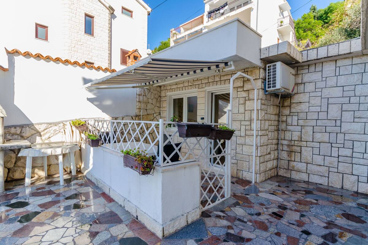 A small outdoor seating area with a white table and chairs, a white pergola with a striped awning, and a white lattice fence with potted plants.