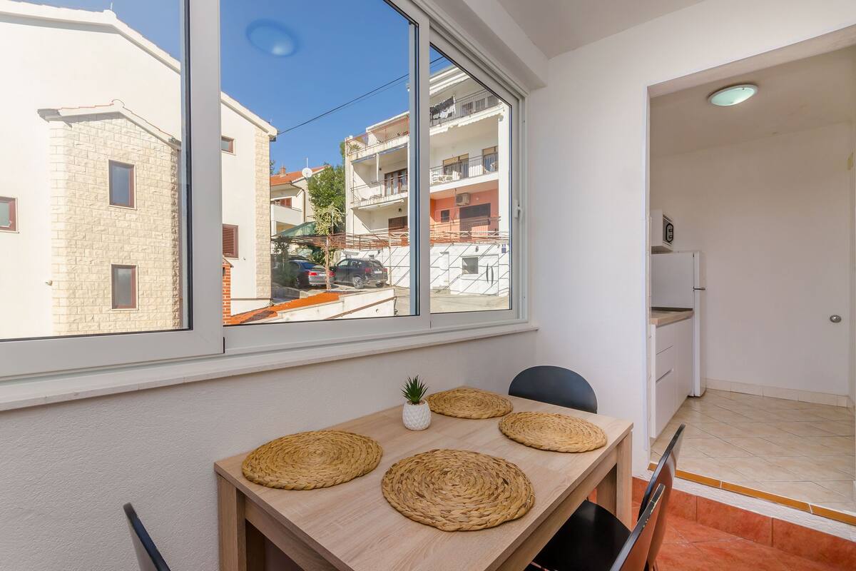 A room with a large window, a wooden table with woven placemats, and a view of neighboring buildings.