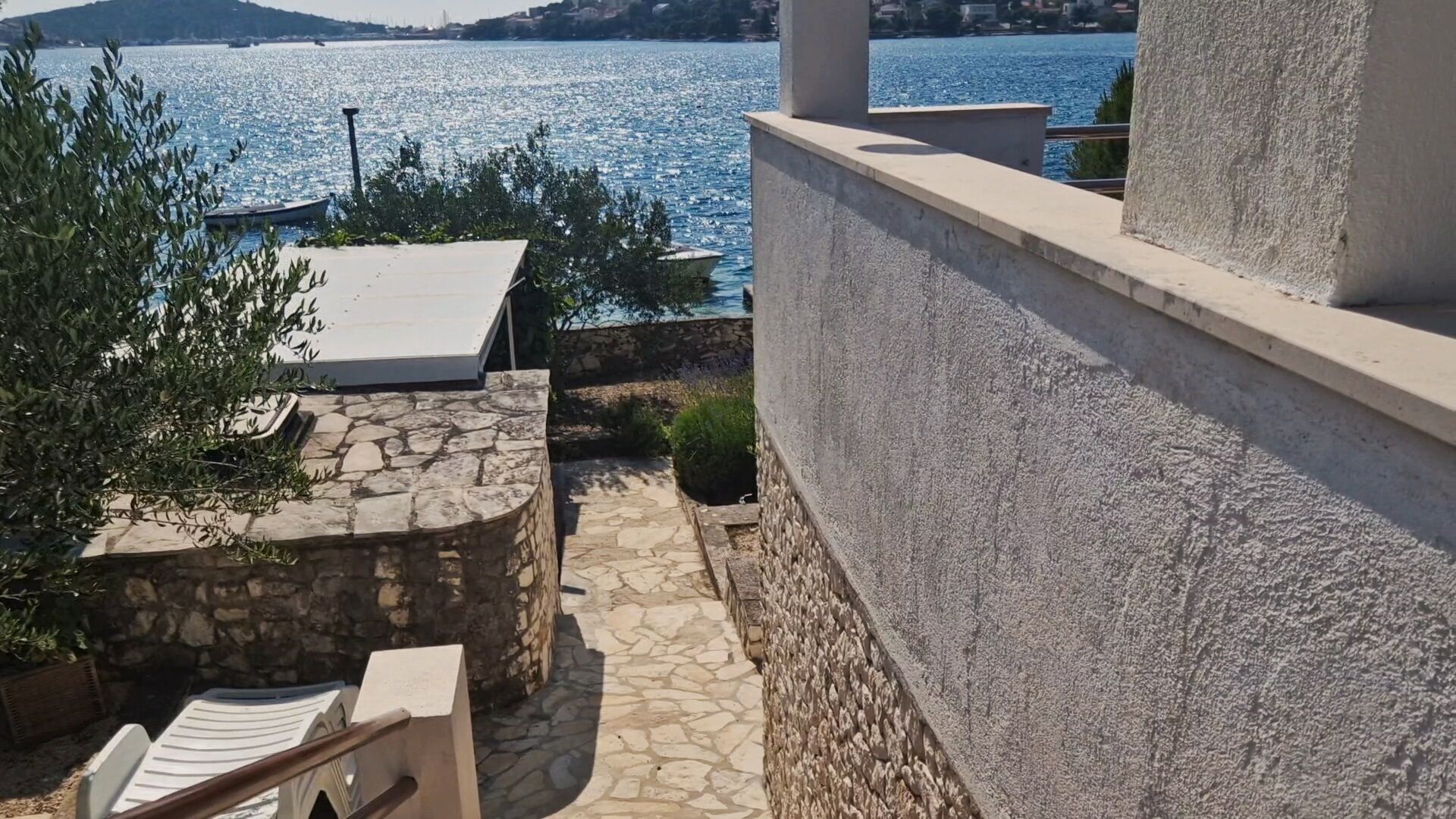 Un camino de piedra conduce a una terraza con vistas al mar, un pequeño jardín y una silla de descanso.