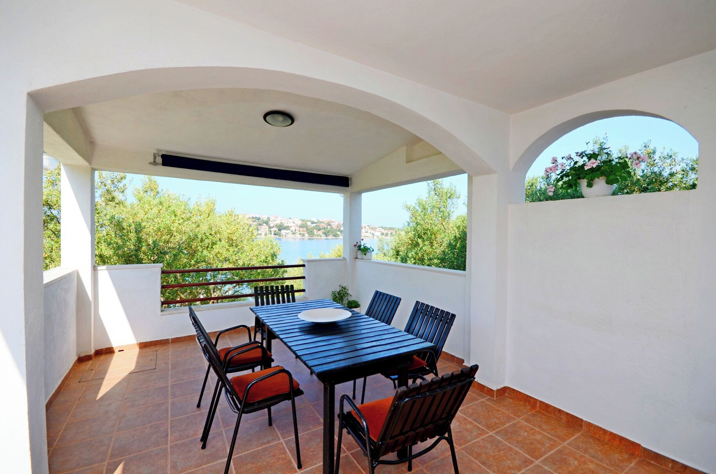 A covered outdoor patio with a table and chairs, a view of greenery and a body of water in the background.