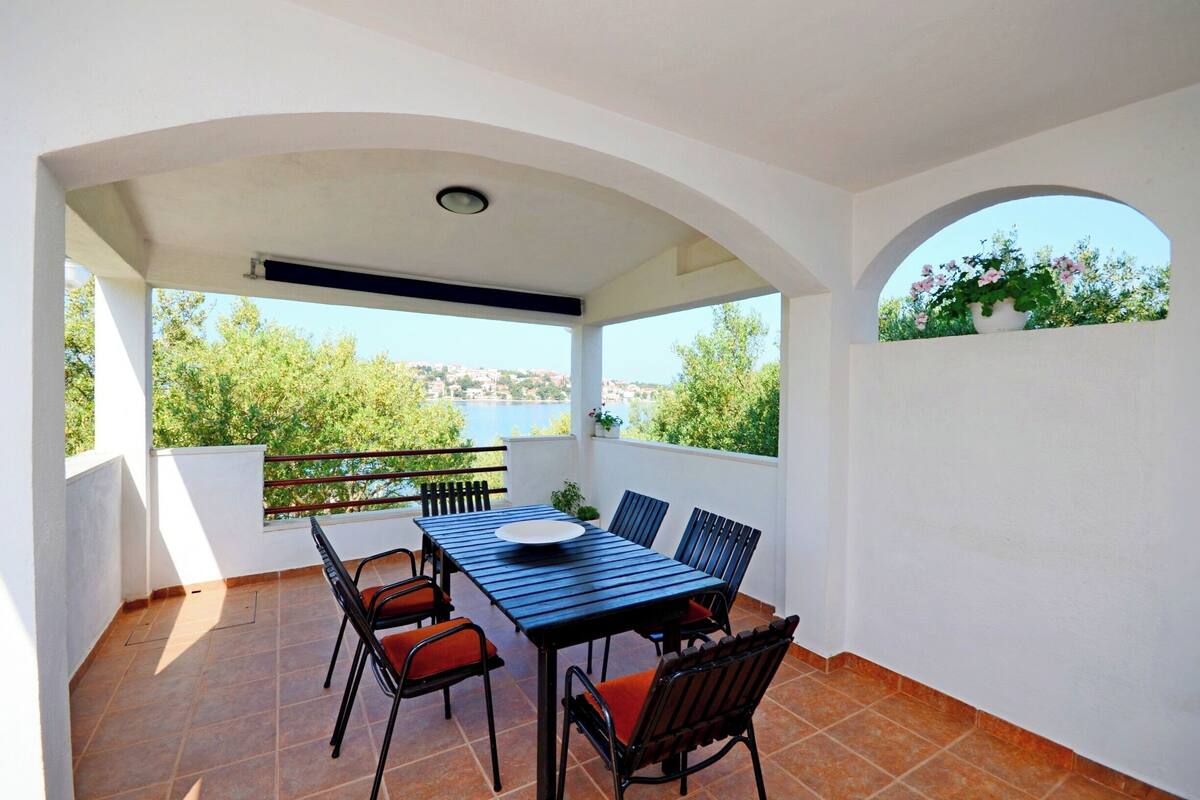 En overdækket terrasse med et bord og stole, udsigt til grøn vegetation og et vandareal i baggrunden.