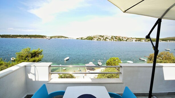 Eine Terrasse mit Tisch und Stühlen, mit Blick auf ein ruhiges Meer mit Booten, einem klaren Himmel und einer entfernten Küstenlinie mit Gebäuden.