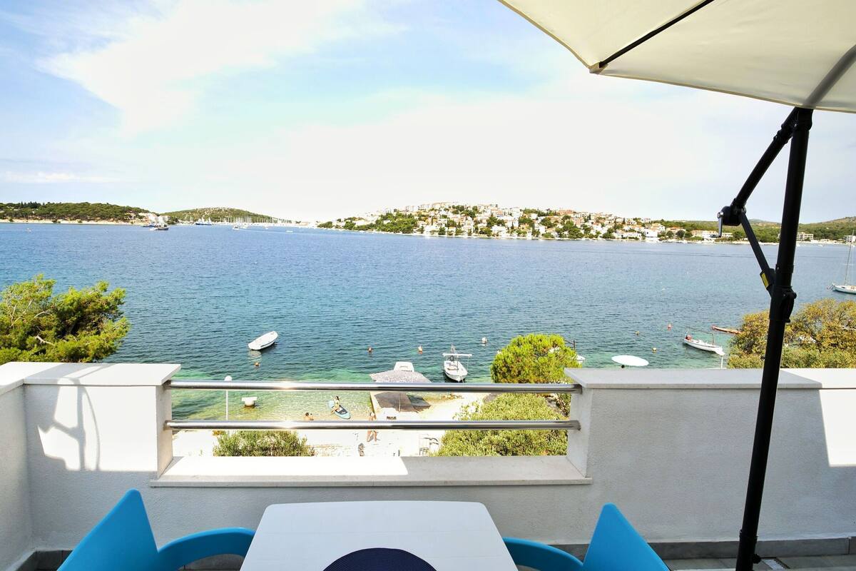 En terrasse med et bord og stole, med udsigt over et roligt hav med både, en klar himmel og en fjern kystlinje med bygninger.