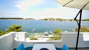 A terrace with a table and chairs, overlooking a calm sea with boats, a clear sky, and a distant shoreline with buildings. - Apartments Jadranka (Rogoznica)