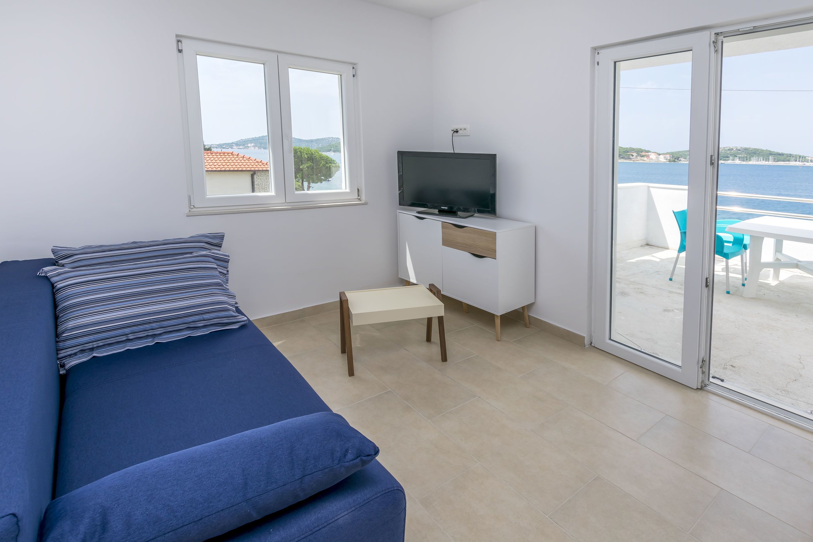 Ein Zimmer mit einem Bett, einem Fernseher auf einem weißen Schrank, einem kleinen Tisch und einer Glastür, die zu einem Außenbereich mit Meerblick führt.