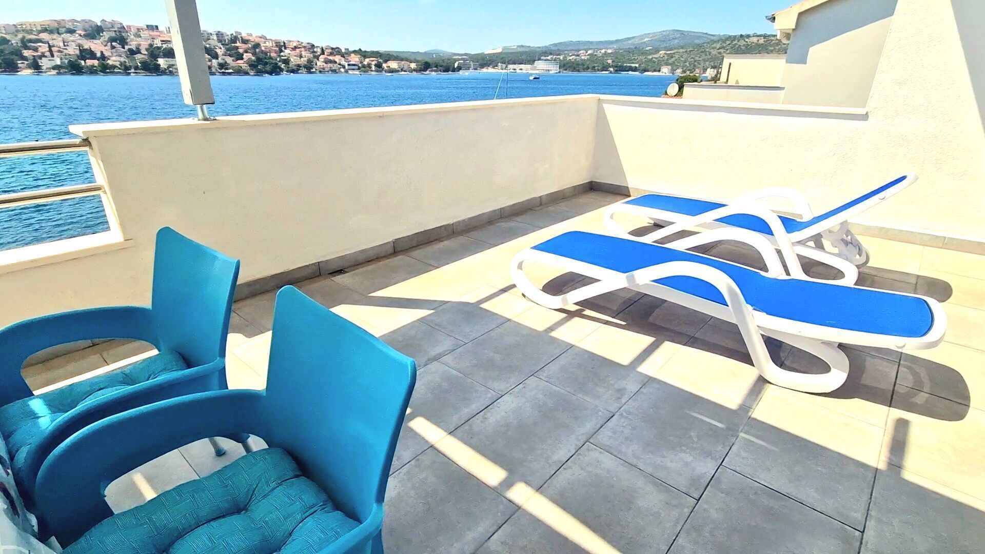 A terrace with blue lounge chairs and a table, overlooking a body of water and a hillside town.