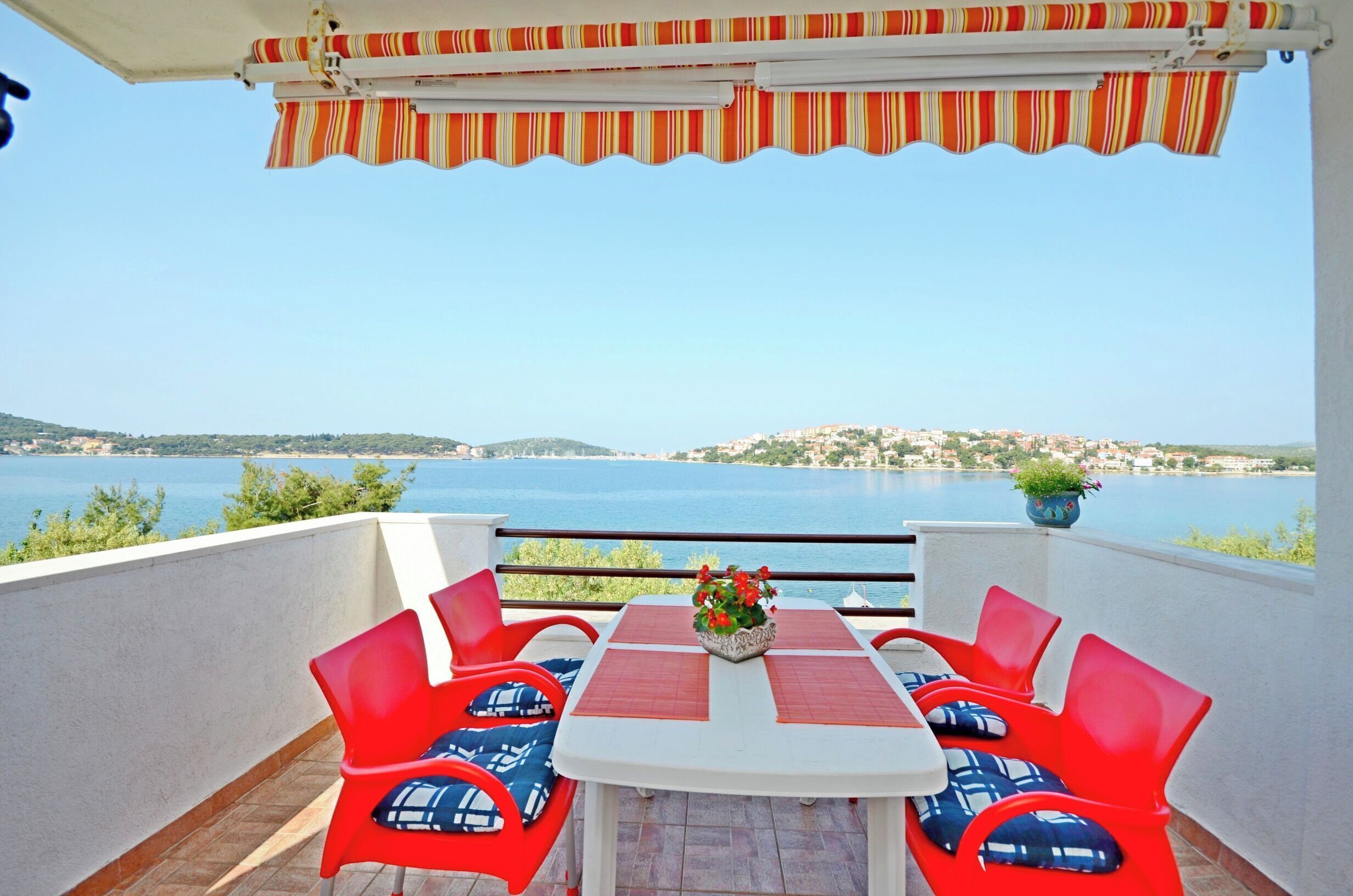 A terrace with a table and red chairs, overlooking a body of water and a distant town.