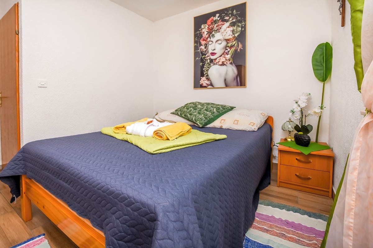 Un letto rifatto con cura, con una coperta azzurra, un cuscino verde e asciugamani piegati. Sopra il letto, un quadro incorniciato, e accanto, un piccolo comodino in legno con una pianta.