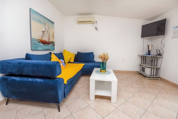 A living room with a blue sofa, a white side table, a painting of a sailboat, an air conditioner, a flat-screen TV, and a small shelf with a microwave.